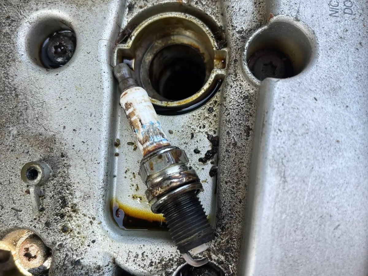 Close-up of a spark plug well with dark oil pooled in and around it, and the old plug lying beside it