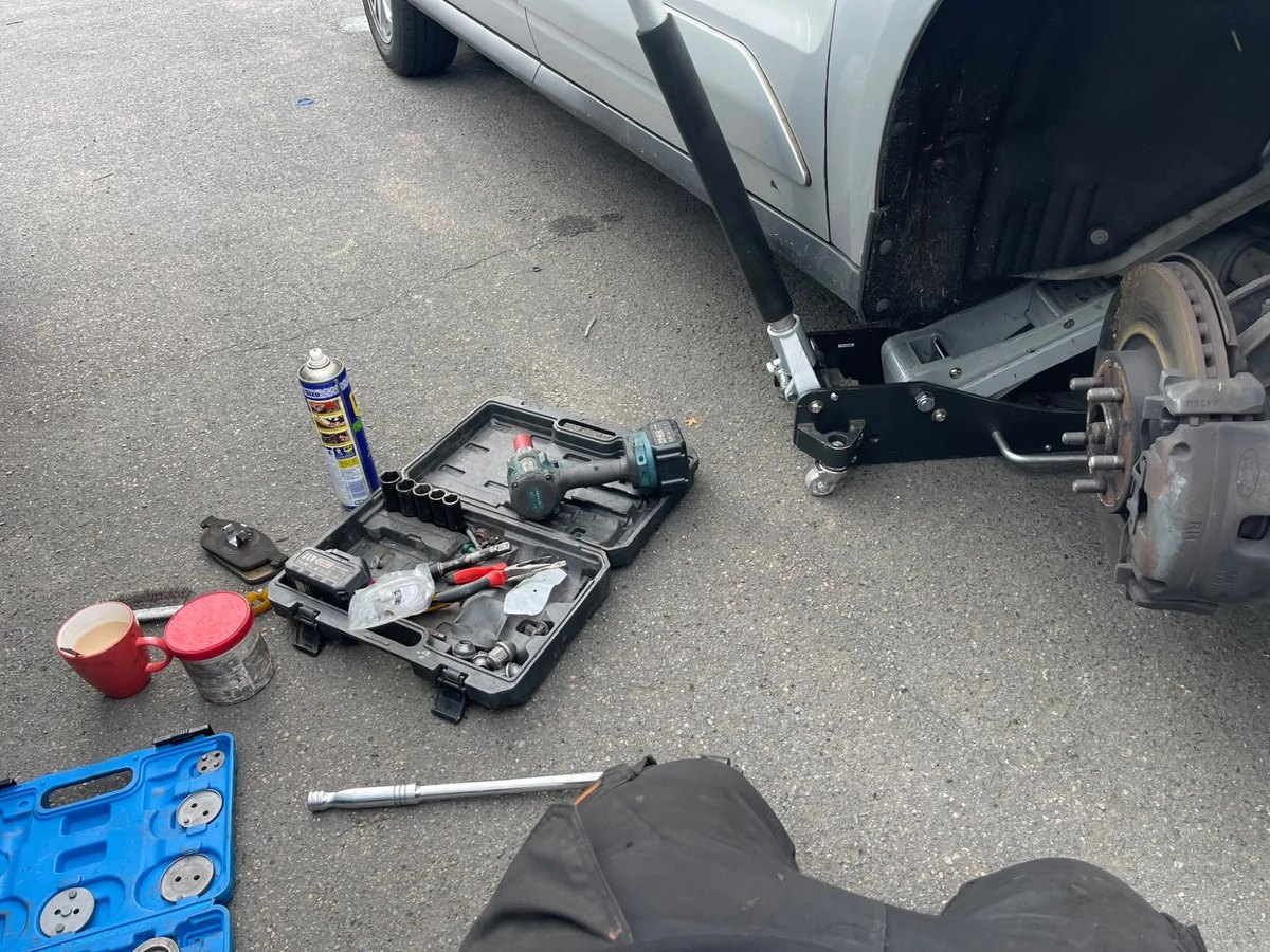 The side of a silver van raised on a trolley jack, with a tool case and a socket set laid out on the ground beside it