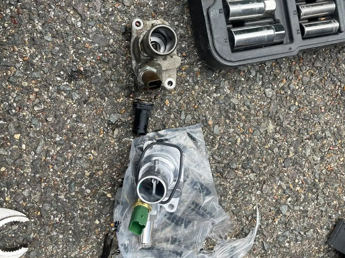 The old thermostat housing on the ground beside the replacement, still sealed in its bag, with a socket set alongside