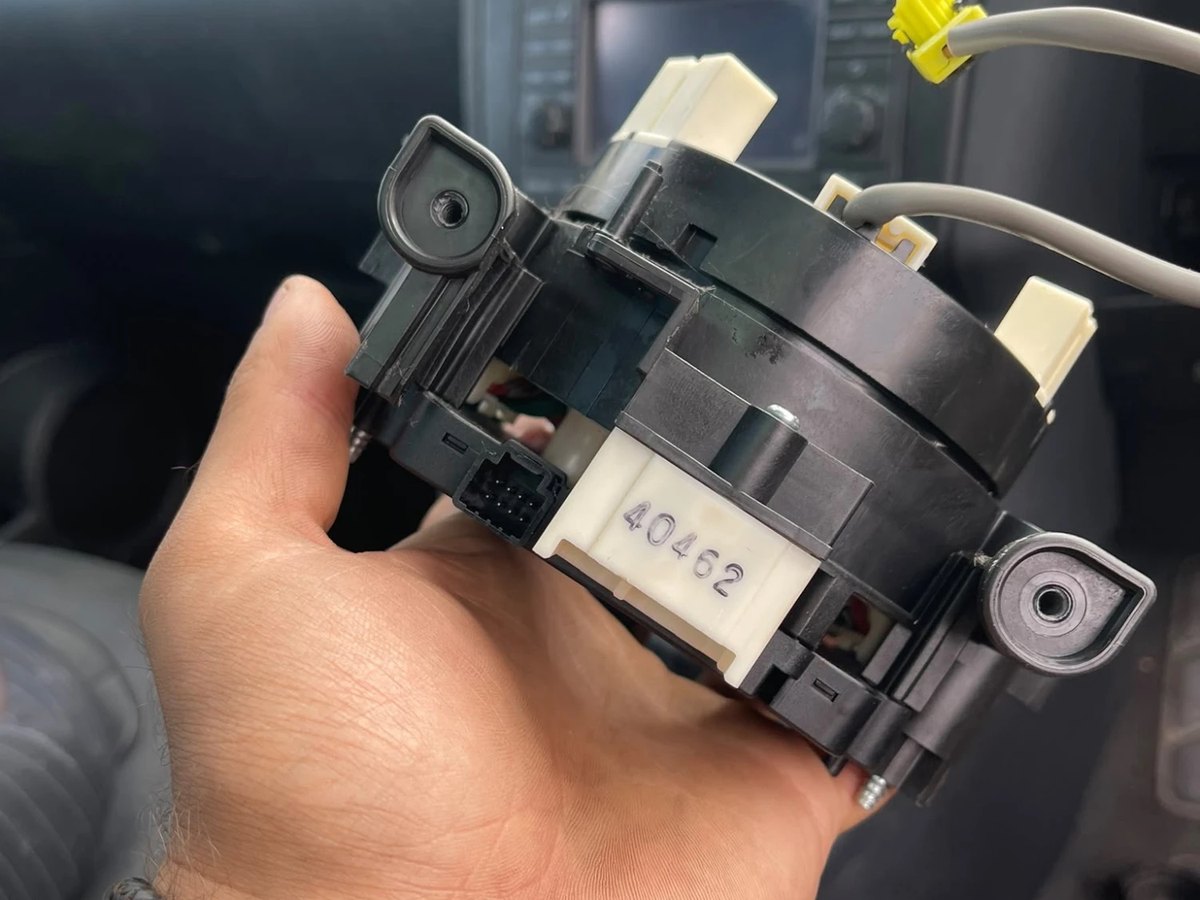 The clock spring unit held in one hand after removal, with its yellow airbag connector and ribbon wiring visible