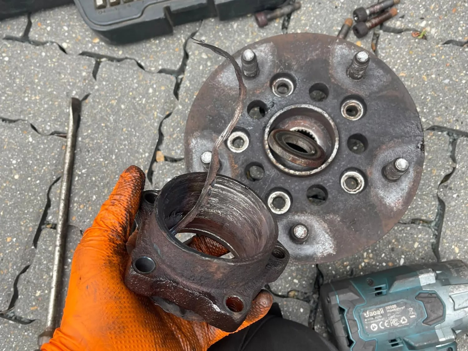 A corroded wheel bearing housing held in a gloved hand, with the old hub flange on the ground behind it